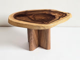 A rustic live-edge wooden table with a thick, polished top showing natural wood grain patterns and a single sturdy leg, set against a clean white background.