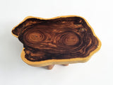 An organic-shaped wooden coffee table made from a live-edge wood slab with a rich dark brown center and golden-yellow outer rim, supported by two small wooden legs on a white background.