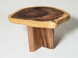 A rustic wooden stool with a thick, live-edge natural wood top and a sturdy, blocky three-legged base, shown on a light gray background.