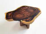A rustic live-edge wooden table with a thick, polished slab top showcasing rich dark brown and golden wood grain patterns, supported by three short wooden legs on a clean white background.