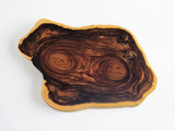 A rustic live-edge wooden serving board or tray with a natural organic shape, showcasing rich dark brown and warm honey-brown wood grain swirls on a light tan edge, photographed on a clean white background.